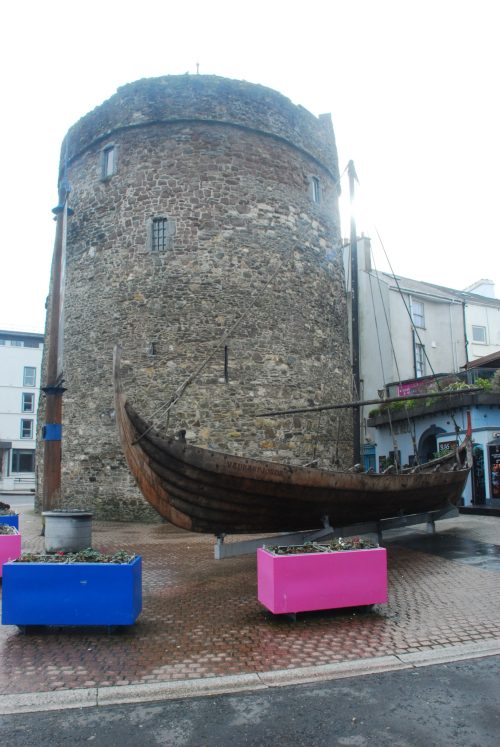 The image shows Reginald's Tower in Waterford, Ireland, a historic stone tower with a rounded top and small windows. In front of the tower, there is a replica of a Viking boat, which is wooden and detailed with ropes and supports. The scene includes colorful planters, one blue and one pink, placed on the cobblestone ground around the boat. The surrounding area includes modern buildings and shops, creating a contrast between the ancient and contemporary elements.