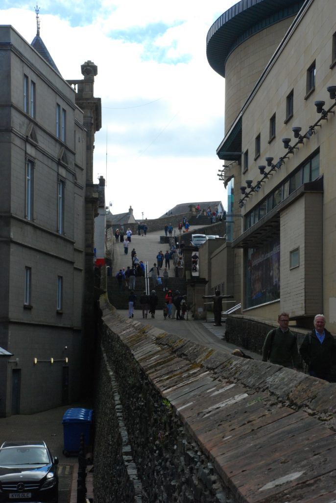 The image shows a section of the Derry Walls in Derry, Northern Ireland. The walls are made of stone and run alongside modern buildings. There are people walking up and down the steps that lead to a higher part of the city. The scene includes a mix of historical architecture and contemporary structures, highlighting the blend of old and new in the city. The sky is partly cloudy, and the area appears to be a popular spot for visitors.