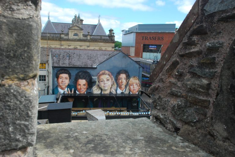 The image features the **Derry Girls mural** in Belfast, Northern Ireland. The mural showcases the five main characters from the hit TV show *Derry Girls*, painted in vibrant colors on the side of a building. The characters—Erin, Orla, Clare, Michelle, and James—are depicted in their school uniforms, with expressive facial features that capture their personalities. The background includes a mix of urban architecture, with nearby buildings and street elements adding to the city atmosphere. The mural is a popular attraction, celebrating the show's cultural impact and humor.