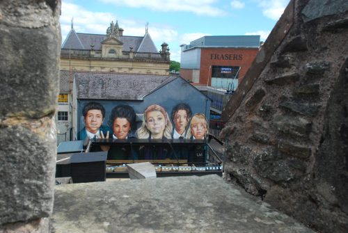 The image features the **Derry Girls mural** in Belfast, Northern Ireland. The mural showcases the five main characters from the hit TV show *Derry Girls*, painted in vibrant colors on the side of a building. The characters—Erin, Orla, Clare, Michelle, and James—are depicted in their school uniforms, with expressive facial features that capture their personalities. The background includes a mix of urban architecture, with nearby buildings and street elements adding to the city atmosphere. The mural is a popular attraction, celebrating the show's cultural impact and humor.