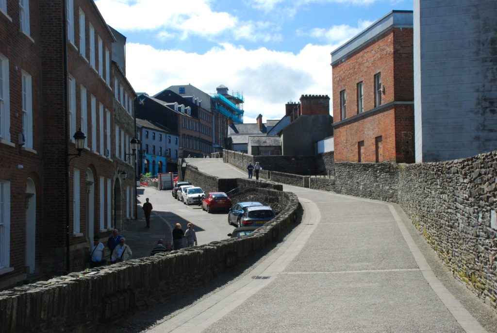 This image shows a section of the Derry Walls in Derry, Northern Ireland. The scene includes a paved walkway along the historic stone walls, with several people walking and cars parked along the adjacent street. The surrounding buildings are a mix of old and new architecture, with brick and stone facades. The sky is partly cloudy, adding to the picturesque view of this historic area.