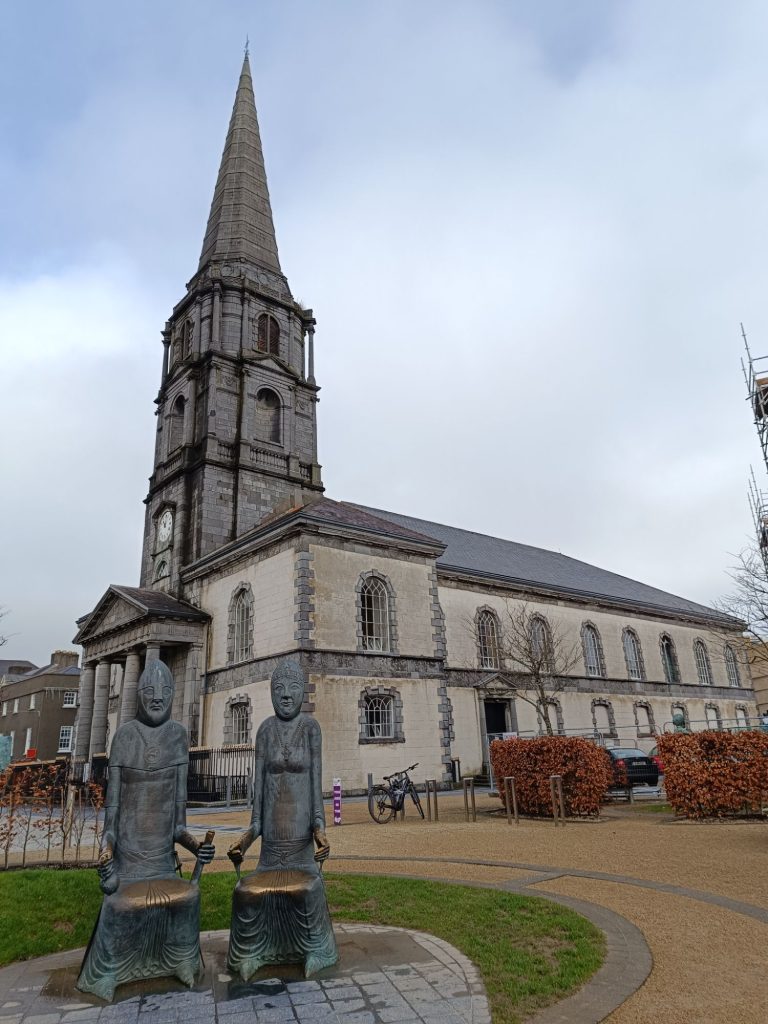 The image shows the exterior of Waterford Christ Church Cathedral, a grand stone building with a tall, pointed spire. In the foreground, two sculptures depict Strongbow and Aoife, who were said to be married at a previous church on this site. The figures are seated and holding hands, positioned on a stone platform surrounded by greenery. The cathedral features arched windows, a clock tower, and an elegant façade, with a few bicycles and cars visible nearby, adding a modern contrast to the historical setting.