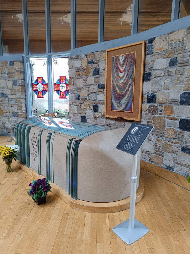 The image shows the tomb of Edmund Rice inside the Edmund Rice Chapel. The tomb is covered with a cloth featuring green and white stripes, and several flowers are placed at its base as a tribute. Behind the tomb, a stone wall holds a framed artwork with a colorful, abstract design. To the left, stained glass windows display red and blue patterns, casting a soft glow into the chapel. A stand with a plaque provides historical context about Edmund Rice. The chapel features wooden flooring and large windows, allowing natural light to fill the space and create a solemn, peaceful atmosphere.