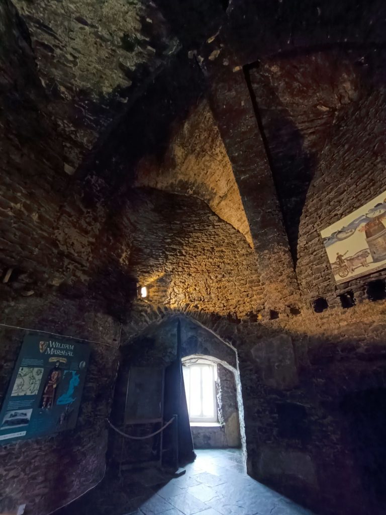 The image shows the interior of Hook Lighthouse in Waterford, Ireland, highlighting its medieval construction methods. The thick stone walls, built using traditional techniques, showcase rough-hewn masonry with curved arches and vaulted ceilings. A window lets in natural light, illuminating the historic craftsmanship. Informational panels on the walls provide context about the lighthouse’s origins, including references to William Marshal, a key figure in its history. The image captures the enduring strength of medieval architecture and the careful preservation of this historic landmark.