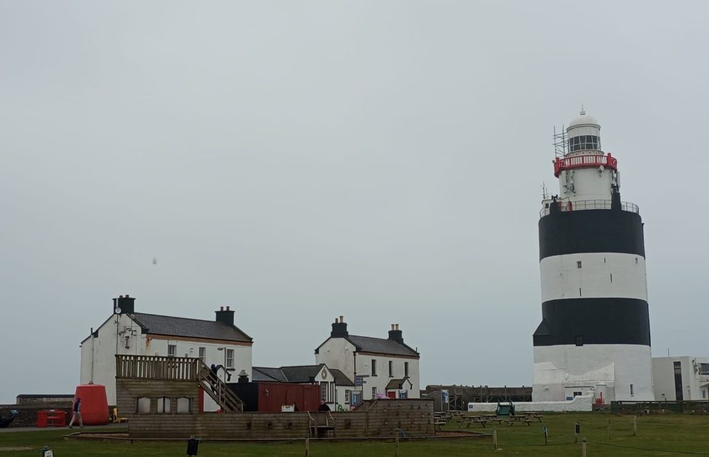 The image shows Hook Lighthouse in Waterford, Ireland, which is the oldest functioning lighthouse in the world. The lighthouse is a tall structure with black and white horizontal stripes and a red railing near the top. Surrounding the lighthouse are several buildings, including two white houses with black chimneys and a wooden deck area. The sky is overcast, and the ground is grassy with some picnic tables and other outdoor furniture.