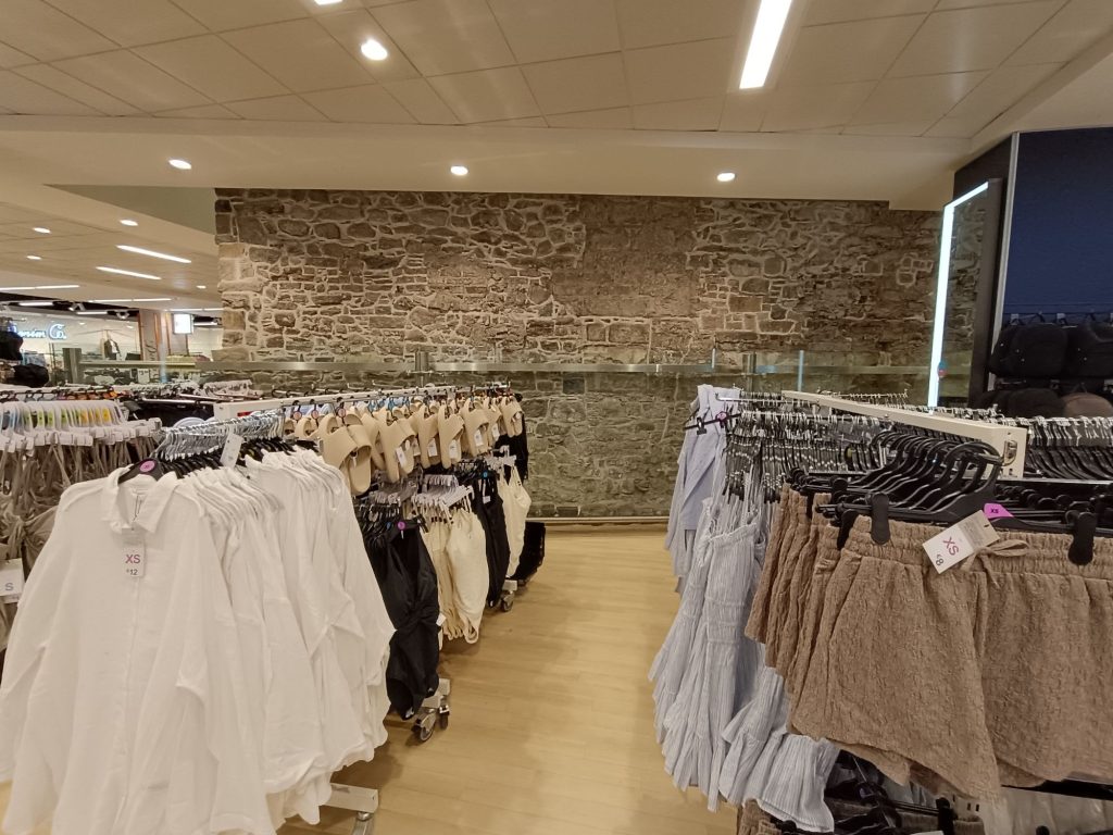 The image shows a section of a medieval stone wall inside a Pennys store in Waterford. The wall is made of irregularly shaped stones and is integrated into the modern retail environment. In front of the wall, there are racks of clothing, including white shirts, beige and black tops, and various shorts. The store has a wooden floor and modern lighting, creating a contrast between the historical wall and the contemporary setting.