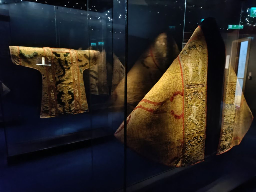 The image shows two medieval vestments displayed in glass cases at the Waterford Medieval Museum. The vestment on the left is a tunic-like garment with intricate embroidery featuring green and black patterns, bordered by red lines. The vestment on the right is a large, triangular-shaped garment with detailed gold embroidery, including depictions of human figures and ornate designs, also bordered by red lines. The dark blue background and reflective glass add to the museum ambiance, highlighting the historical significance and craftsmanship of these garments.
