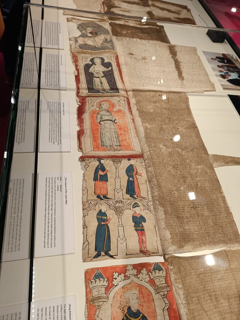 The image shows the Waterford Charter housed in the Medieval Museum. The historic document, displayed in a glass case, consists of several parchment sheets covered in medieval script and colorful illustrations. These illustrations depict figures such as knights and clergy, dressed in period attire. Nearby informational panels offer historical context about the charter’s significance in medieval governance. The careful preservation of the document highlights its importance in Ireland's history.