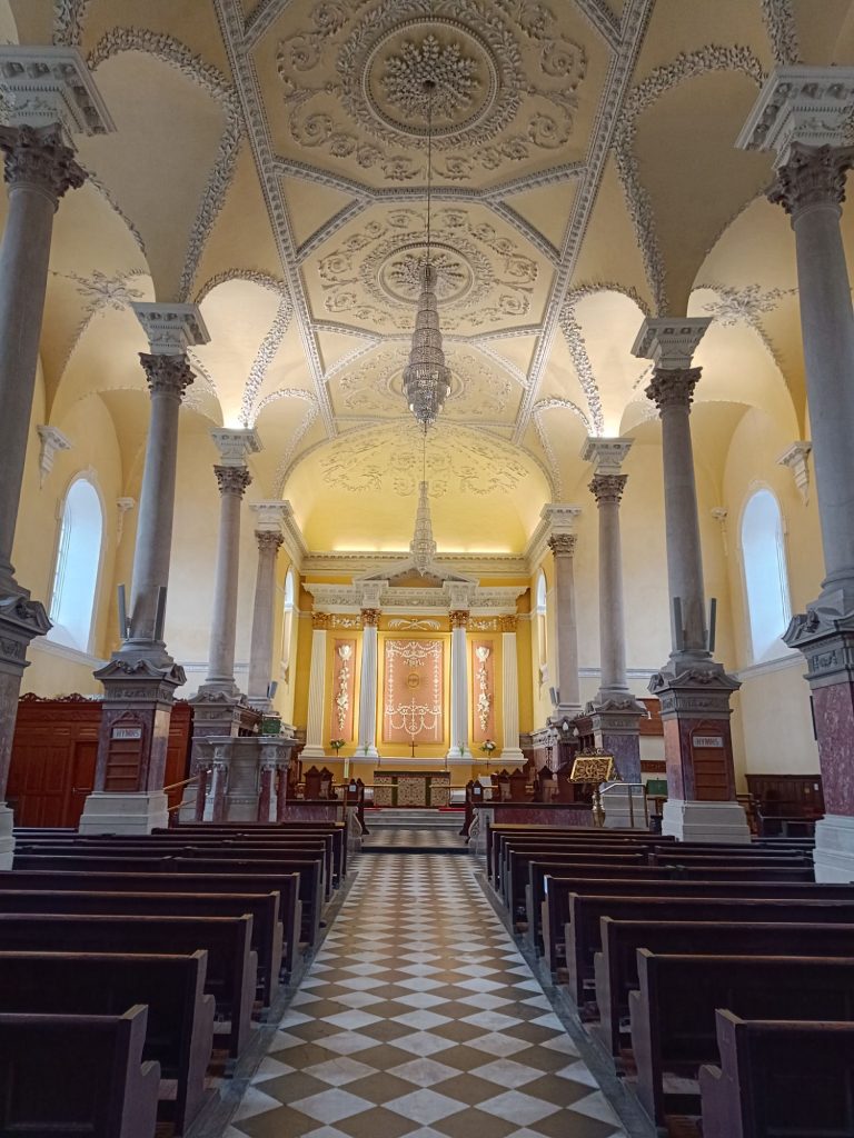 The image shows the interior of a historic Christ Church Cathedralin Waterford, Ireland, where Strongbow and Aoife were married. The church features a grand vaulted ceiling with elegant architectural details. Tall columns line the sides, supporting the structure, while rows of wooden pews lead up to a beautifully adorned altar. A striking chandelier hangs in the center, adding to the solemn yet majestic atmosphere of this significant historical site.