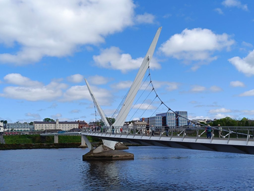 The image shows the **Peace Bridge in Derry, Northern Ireland**, a modern **cycle and footbridge** spanning the **River Foyle**. The bridge features **two large, angled supports** with cables creating a striking visual design. Several **people are walking across the bridge**, enjoying the scenic view. In the background, a mix of **older and newer buildings** lines the riverbank. The sky is **blue with scattered clouds**, adding to the bright and inviting atmosphere. The bridge is a **symbol of peace and unity**, connecting different parts of the city.