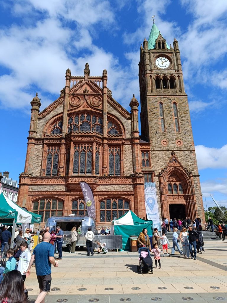 The image shows the **Guildhall in Derry**, a historic building with a striking **red sandstone facade** and a **tall clock tower**. The Guildhall features intricate **Gothic-style architecture**, with **large stained glass windows** and detailed carvings. In front of the building, several **green and white tents** are set up, indicating a **market or event** taking place. People are walking around and interacting with the stalls, creating a **lively atmosphere**. The sky is **blue with scattered clouds**, adding to the vibrant and inviting scene.