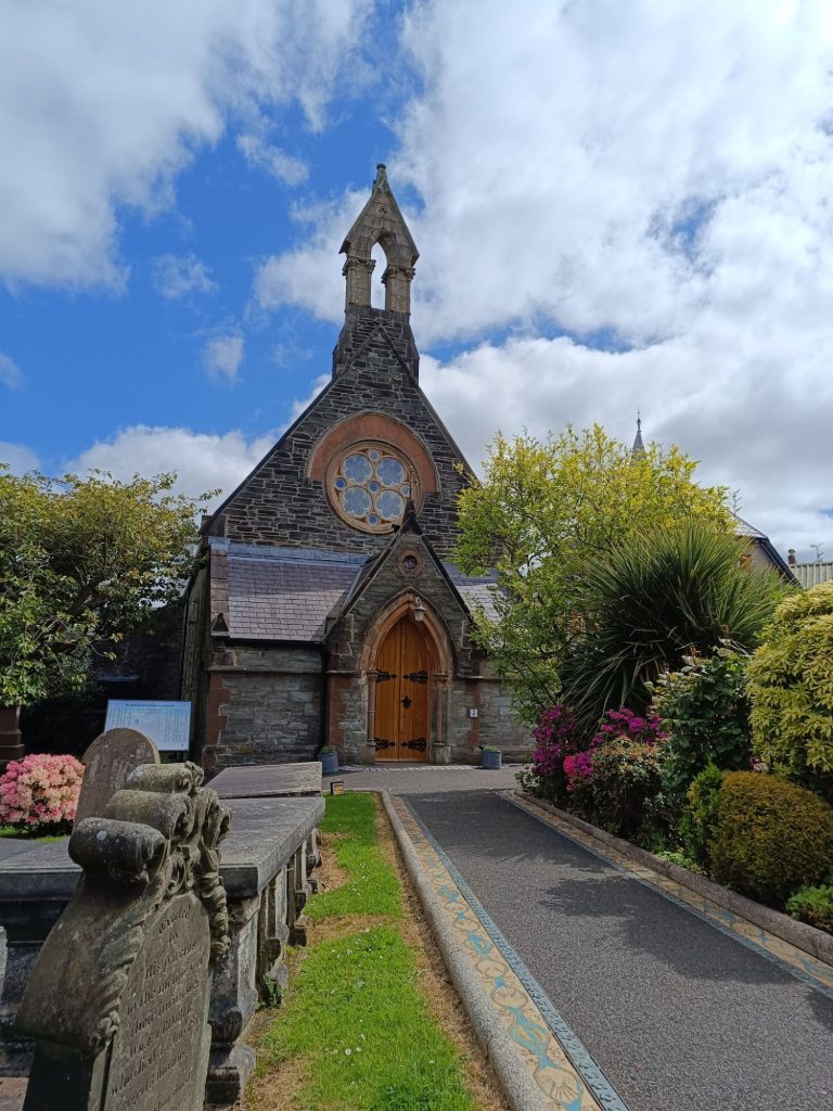 The image shows **St. Augustine's Church**, also known as the **Wee Church on the Walls**, in Derry, Northern Ireland. The church has a **stone facade** with a **pointed bell tower** and a **large circular stained glass window** above the entrance. The **wooden double doors** lead into the historic building, which is surrounded by **well-maintained gardens** with various plants and flowers. A **gravestone** is visible on the left side, suggesting an adjoining cemetery. The sky is **partly cloudy**, with patches of blue visible, adding to the serene atmosphere of this historic site.